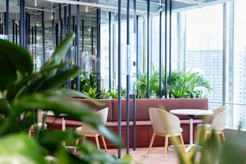 Modern office lounge with pink cushioned seating, round white tables, beige chairs, large windows, and green plants in the foreground.