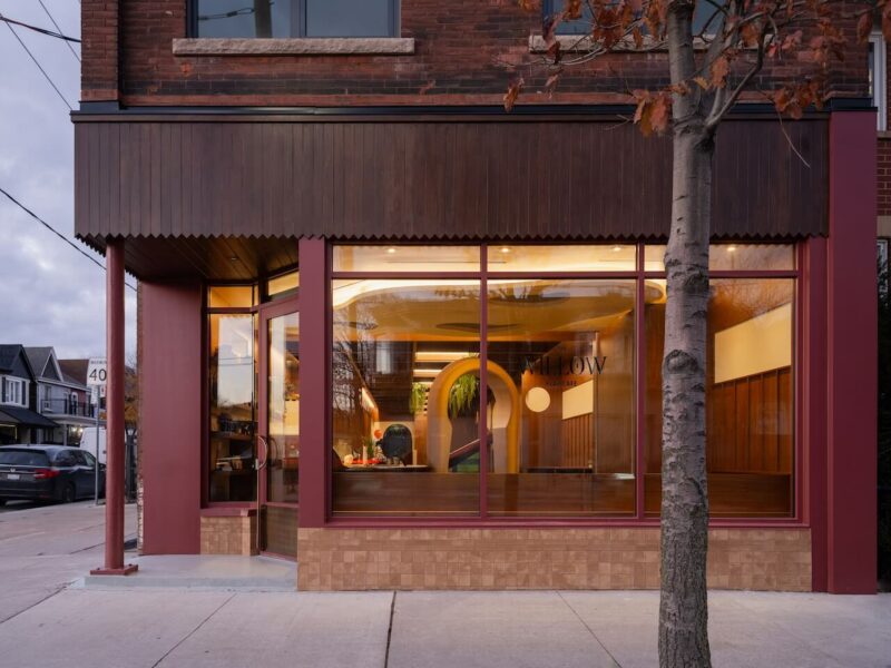 A street view of a modern storefront with large windows, warm interior lighting, and a tree in front on the sidewalk.