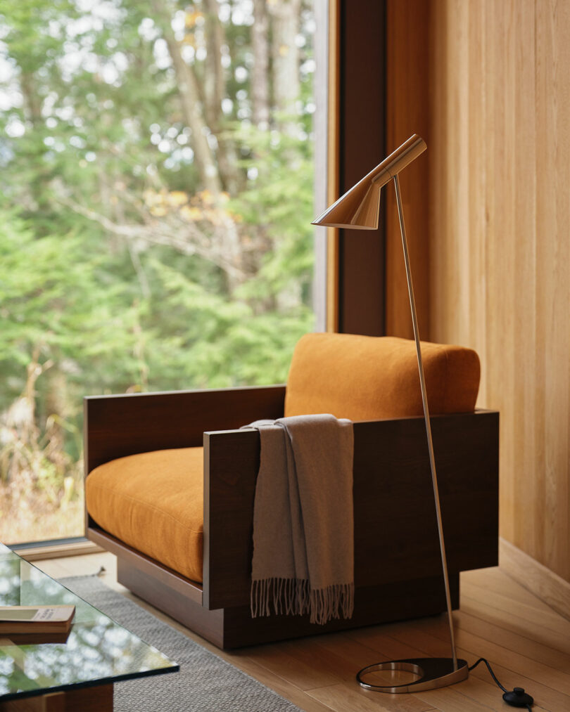 A modern Design Within Reach armchair with an orange cushion and a gray throw sits beside a floor lamp and large window overlooking green trees.