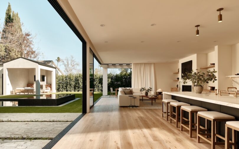 Modern open-concept living space with a kitchen island, stools, and a living area, opening to a patio and backyard with a small outdoor lounge structure.