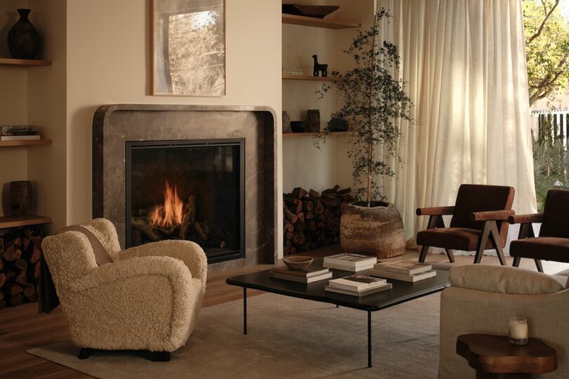 A cozy living room features a lit fireplace, a plush armchair, wooden chairs, a coffee table with books, and a potted tree near large curtained windows.