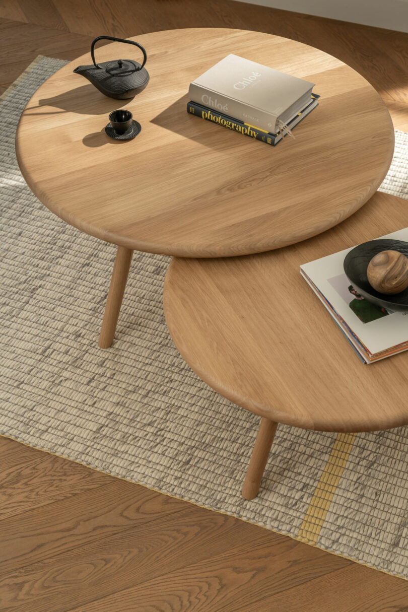 Two round wooden coffee tables on a woven rug, each with books, a black teapot, and a small black cup arranged on top.
