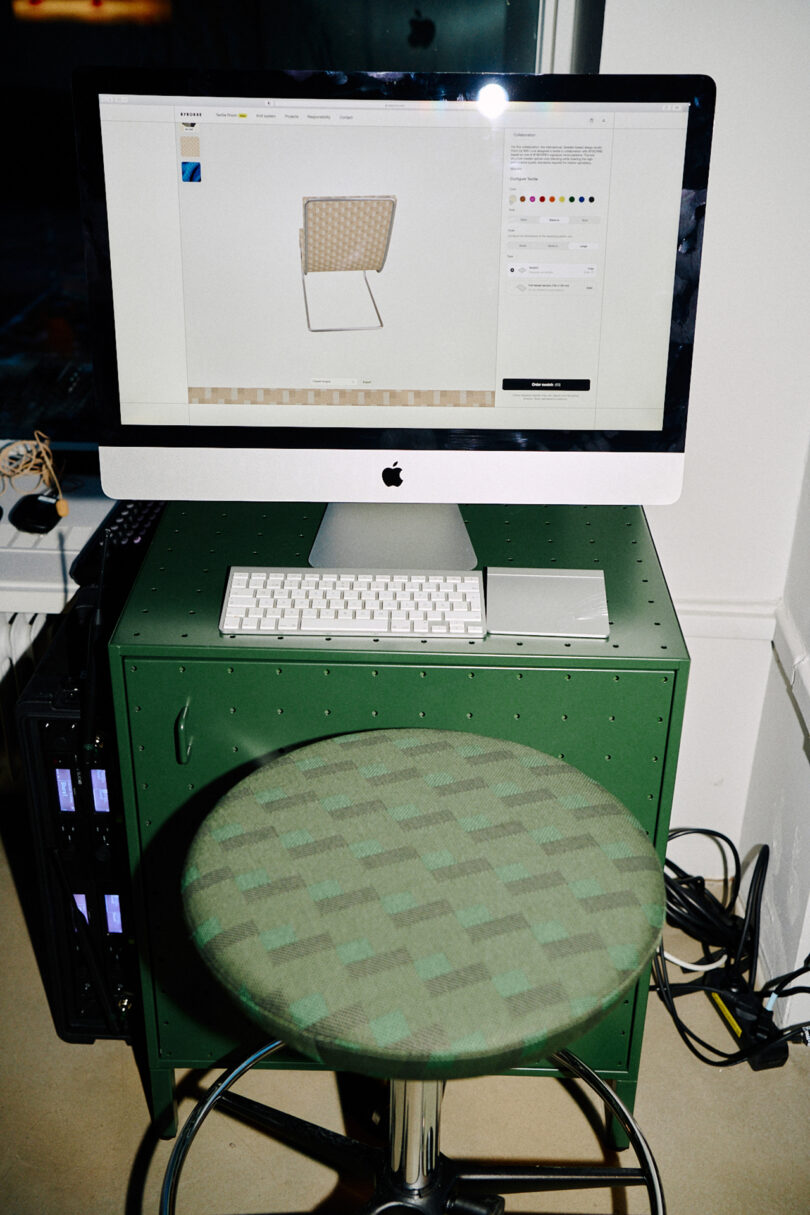 An iMac displaying a 3D modeling program sits on a green cabinet, alongside a white keyboard and mouse. A green checkered stool inspired by Form Us With Love design sensibilities stands in front, with cables visible on the floor.
