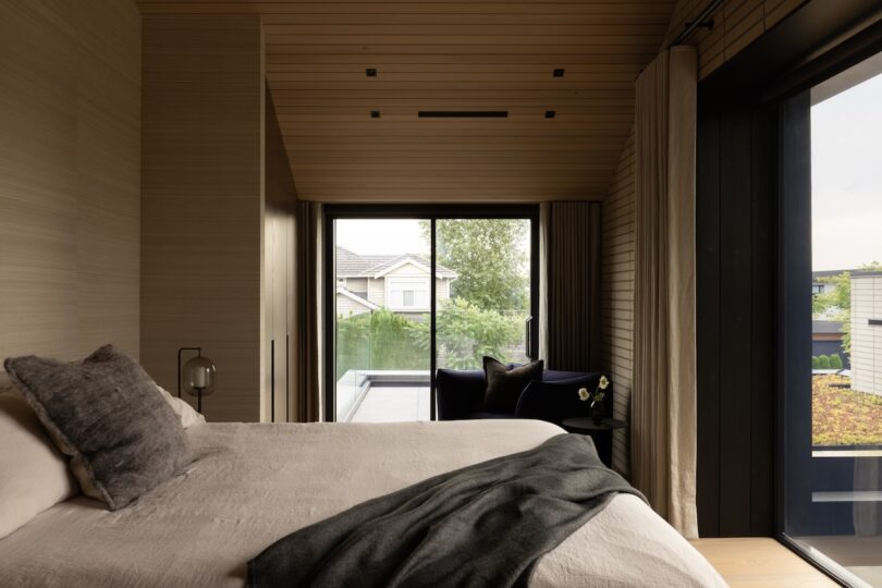 Modern bedroom with neutral tones, large window, and sliding glass door opening to a view of houses and greenery outside. Gray bedding and a cushioned chair are visible.