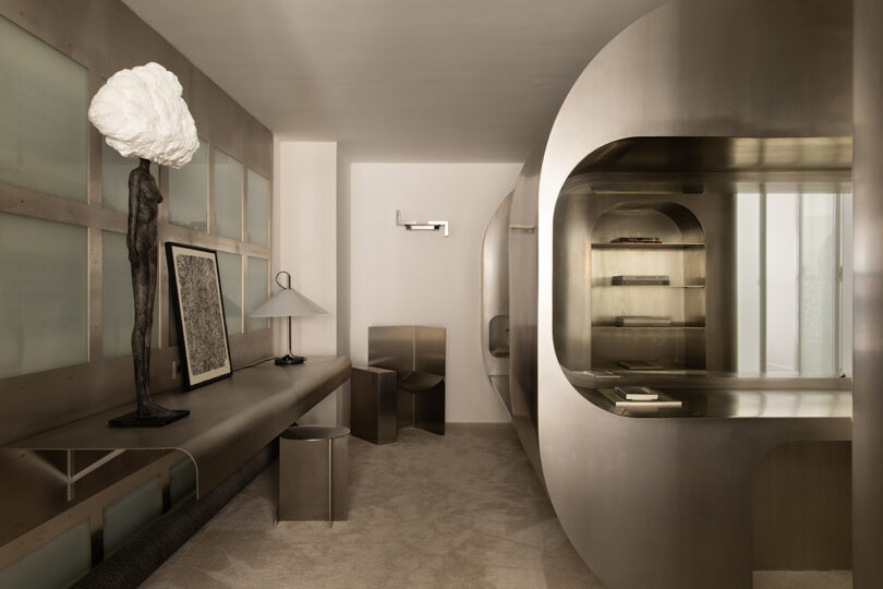 Modern minimalist room with metallic furniture, abstract sculpture lamp, a framed artwork, curved shelving unit, and neutral-toned walls and flooring.