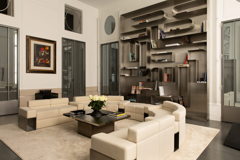 Modern living room with cream modular sofas, a square coffee table with a vase of white flowers, a geometric metal bookshelf with books and decor, and a framed painting on the wall.