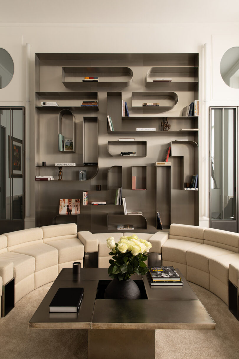 Modern living room with a large, curved metal bookshelf holding books and decor, beige sectional sofas, a dark coffee table, and a vase of white flowers in the center.