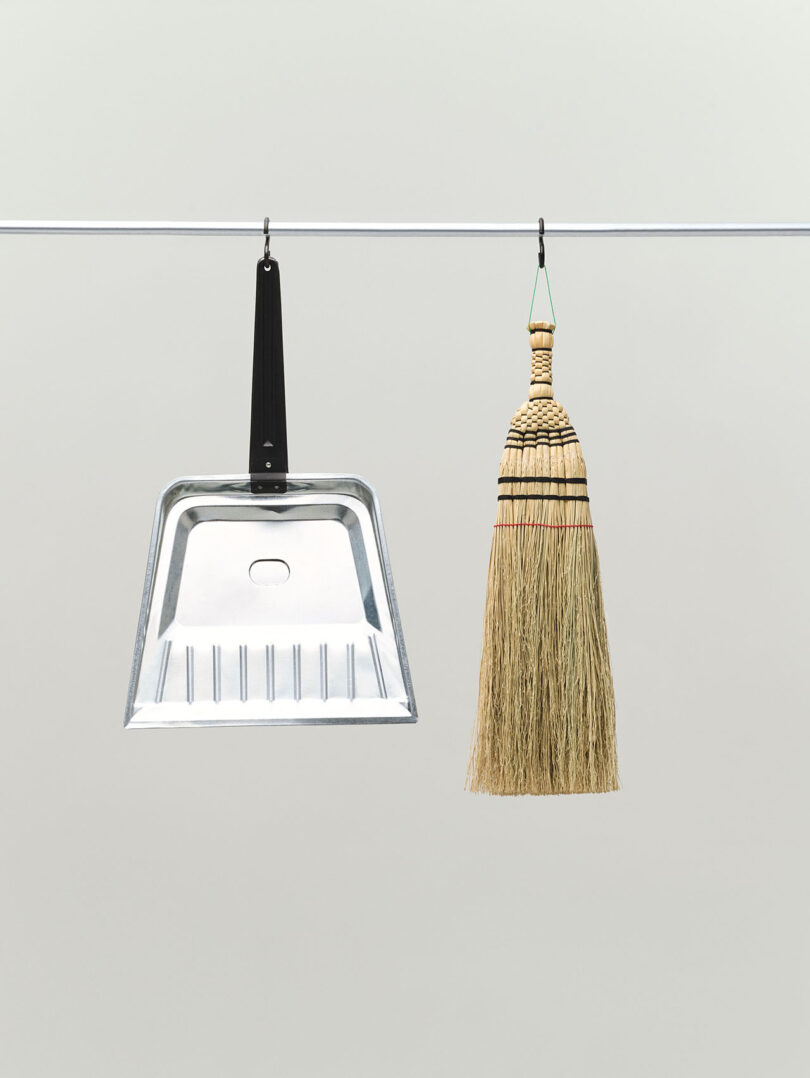 A metal dustpan and a straw broom hanging side by side on a metal rod against a plain background.