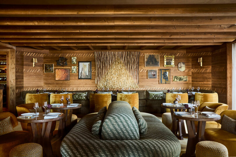 A cozy restaurant interior with wooden walls, eclectic wall art, round tables set with plates and glasses, and plush green and gold seating.