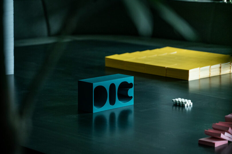 A teal rectangular object with geometric cutouts sits on a dark surface, near stacked yellow papers and small white objects, with pink items visible in the corner.