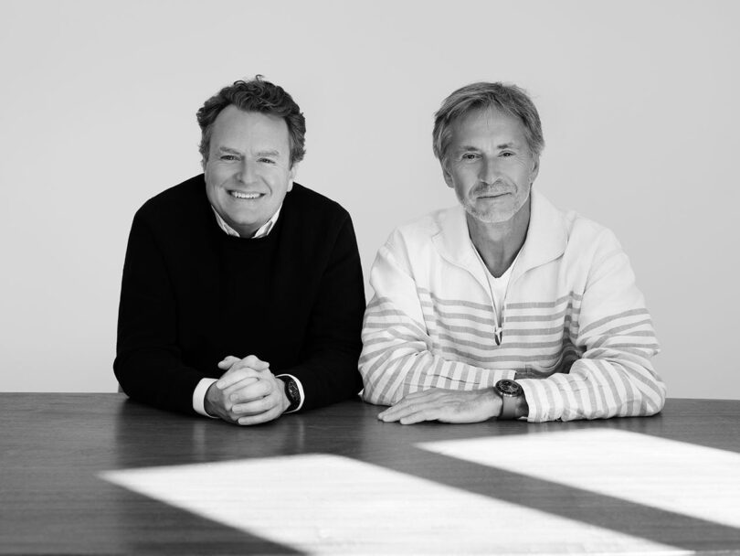 Two men sit side by side at a table, facing the camera and smiling, in a well-lit room with a plain TYPE 3 background.