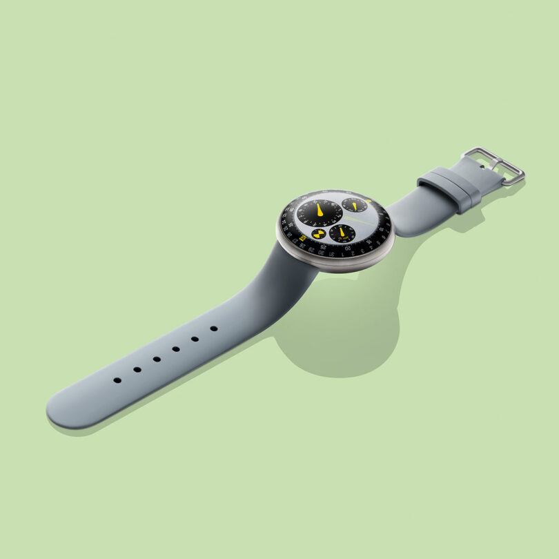 A modern TYPE 3 wristwatch with a gray strap and a round face displaying three yellow and black dials, set against a light green background.