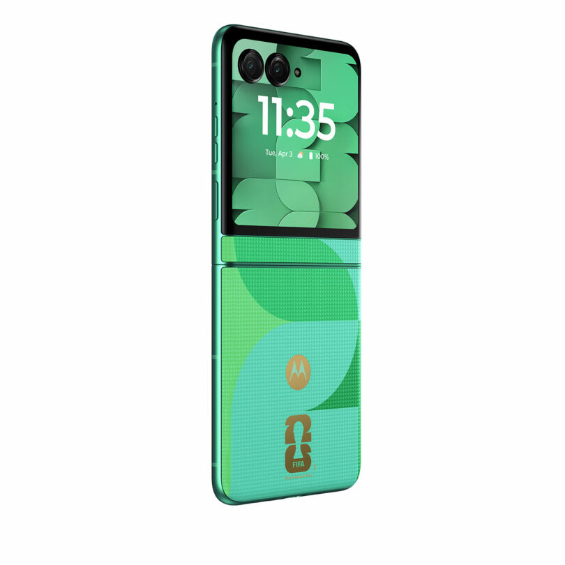 A green Motorola flip phone is shown partially open, featuring dual rear cameras, a patterned exterior, and a FIFA logo on the lower back panel.