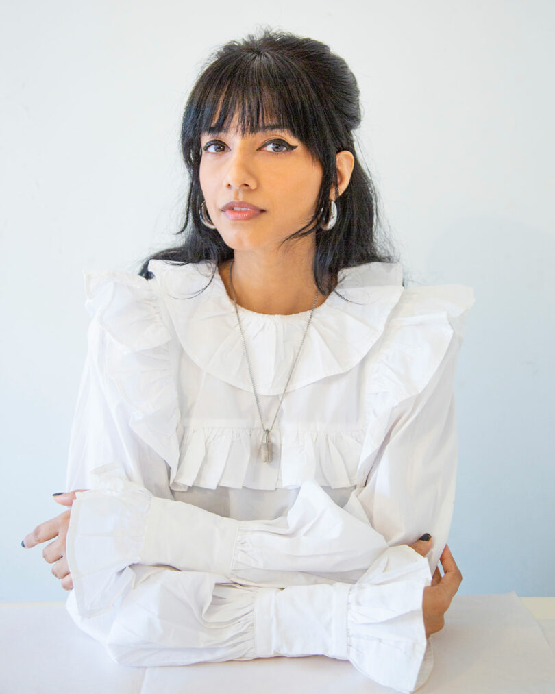 A woman with long dark hair and bangs, wearing a white ruffled blouse and a necklace, sits at a table with her arms crossed against a plain light background.