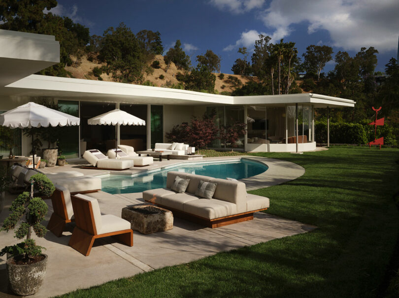 Modern OSKLO house with large glass windows, a swimming pool, lounge chairs, umbrellas, and manicured lawn surrounded by trees and hills under a partly cloudy sky.