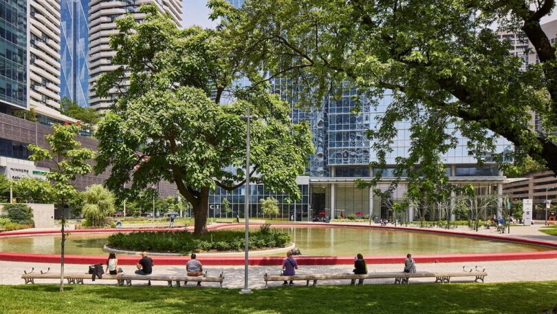 People sit on benches facing a circular pond in an urban park with tall buildings and trees in the background on a sunny day.