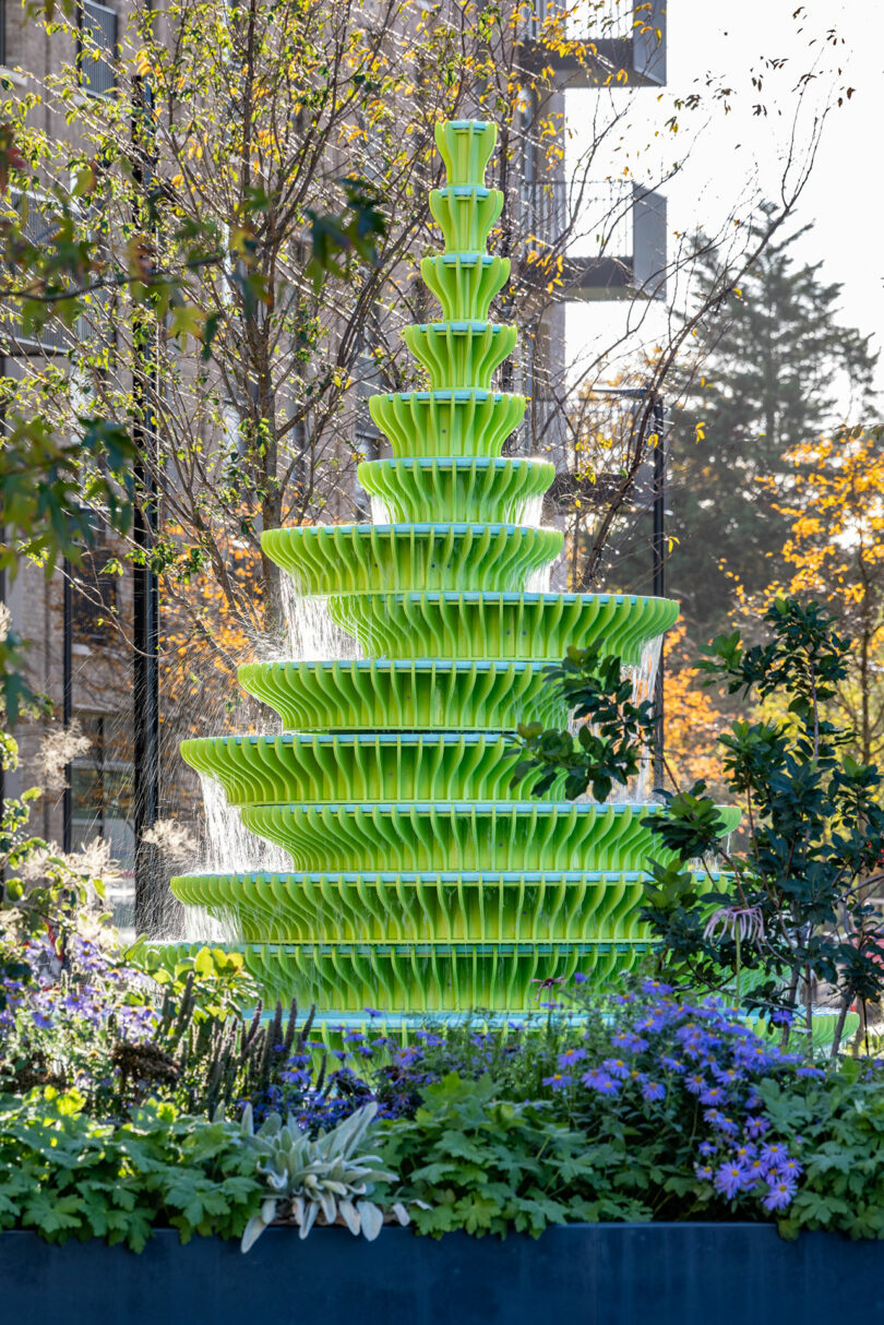 A tiered, bright green modern fountain stands in a garden with surrounding plants and flowers, with water cascading down its layers. Trees and a building are in the background.