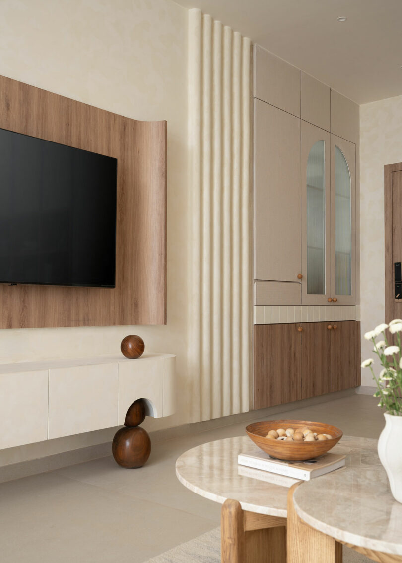 Modern living room with wall-mounted TV, floating cabinet, wood accents, decorative spheres, and a round coffee table with a bowl and vase of flowers. Built-in storage has arched glass doors.
