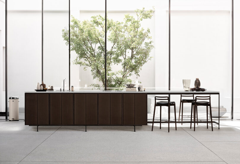 Modern kitchen with a Vipp dark wood island, minimalist decor, three barstools, and large windows showcasing a green tree outside.